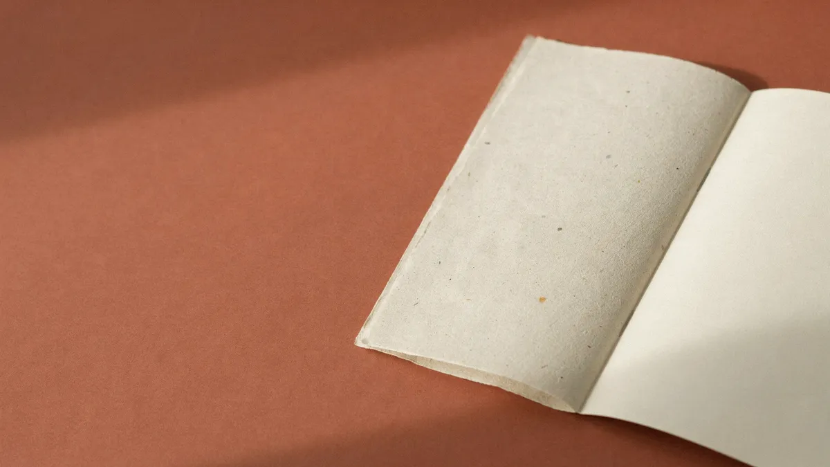 Gefaltetes Blatt Papier auf mattem Terrakotta, am Bildrand angeschnitten, eine Hälfte mit sichtbarer Papierfaser, die andere glatt und makellos.